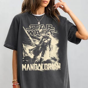 Pu&ograve; includere: T-shirt grigio scuro con grafica invecchiata di The Mandalorian di Star Wars. Il design include il logo di Star Wars e la parola "Mandalorian". La maglietta ha maniche corte e girocollo.