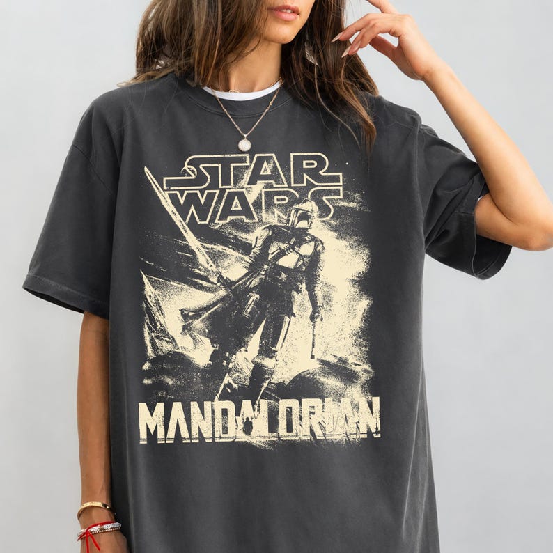 Pu&ograve; includere: T-shirt grigio scuro con grafica invecchiata di The Mandalorian di Star Wars. Il design include il logo di Star Wars e la parola "Mandalorian". La maglietta ha maniche corte e girocollo.