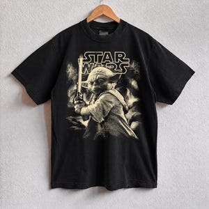 Może przedstawiać: Czarny t-shirt Star Wars z sepiowym wizerunkiem Yody trzymającego miecz świetlny. Logo Star Wars znajduje się nad obrazem. T-shirt wisi na drewnianym wieszaku.
