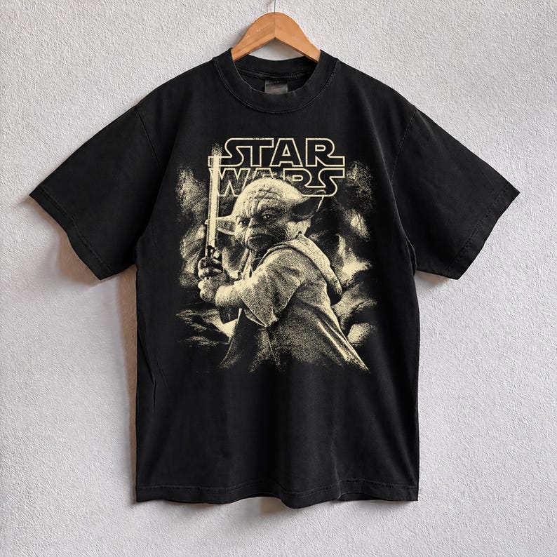 Może przedstawiać: Czarny t-shirt Star Wars z sepiowym wizerunkiem Yody trzymającego miecz świetlny. Logo Star Wars znajduje się nad obrazem. T-shirt wisi na drewnianym wieszaku.