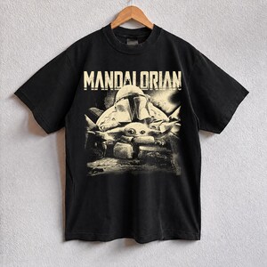 May include: Black t-shirt with a distressed graphic of The Mandalorian and Grogu. The word "MANDALORIAN" is printed in white across the top. The design has a vintage, worn look.