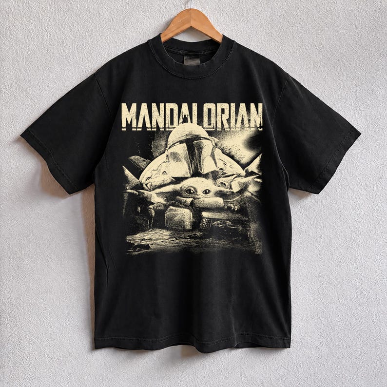 May include: Black t-shirt with a distressed graphic of The Mandalorian and Grogu. The word "MANDALORIAN" is printed in white across the top. The design has a vintage, worn look.