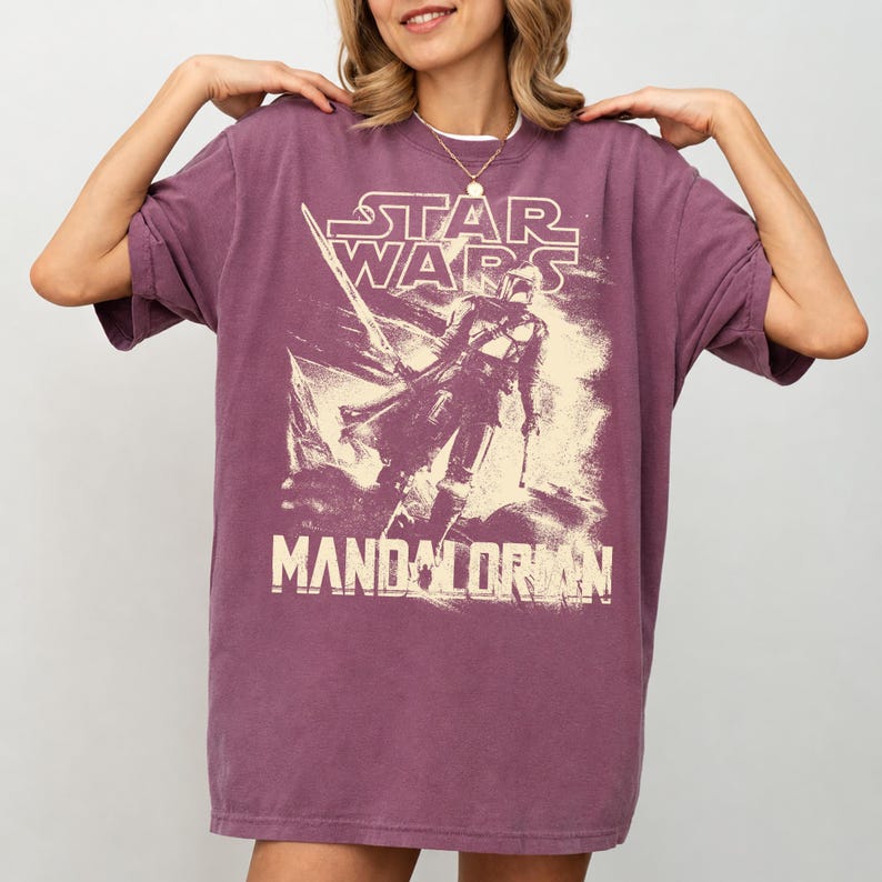 Pu&ograve; includere: Una maglietta color malva con una grafica di Star Wars. Il design include le parole "STAR WARS" sopra un'illustrazione di The Mandalorian, con la parola "MANDALORIAN" sotto. La maglietta ha maniche corte e una vestibilit&agrave; ampia.