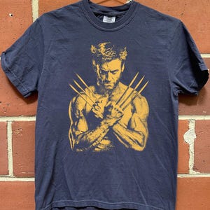 Peut inclure: Un t-shirt bleu marine à manches courtes avec une impression graphique jaune représentant un personnage musclé aux trois griffes acérées étendues de chaque main, croisées sur la poitrine. Le personnage a un regard concentré.