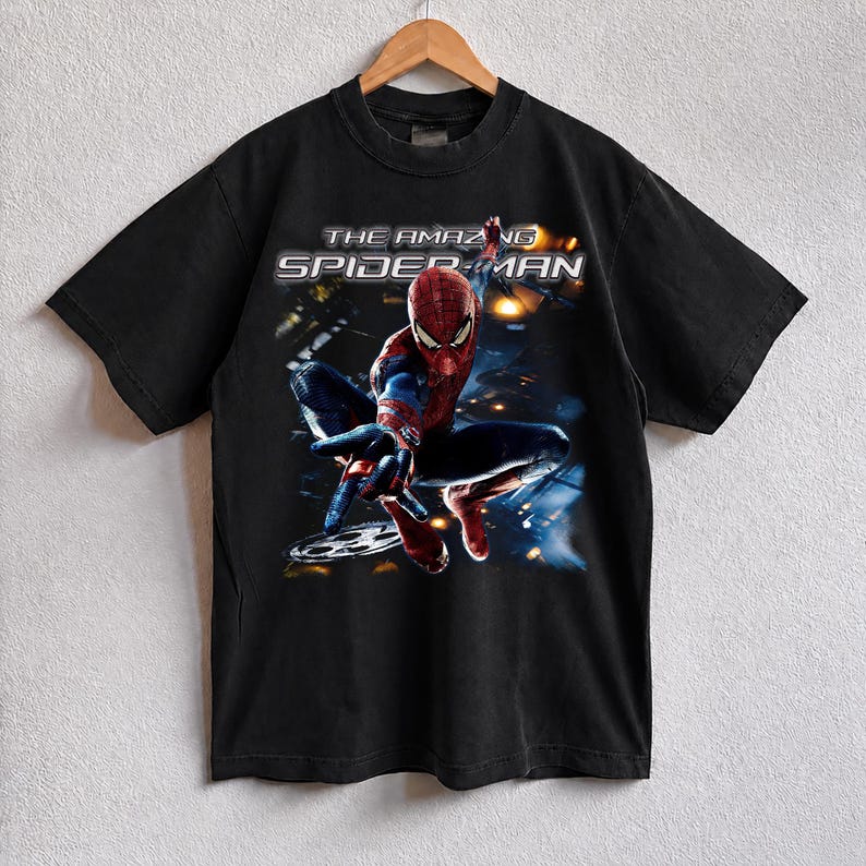 May include: Black t-shirt featuring a graphic of Spider-Man in a dynamic pose. The superhero is depicted in his classic red and blue suit, with the words "THE AMAZING SPIDER-MAN" printed above him. The shirt is hanging on a wooden hanger.