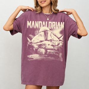 May include: A purple t-shirt with the word "MANDALORIAN" printed across the top. Below the text is a graphic of a Mandalorian helmet and a small creature. The shirt has short sleeves and a loose fit.