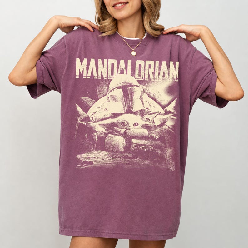 May include: A purple t-shirt with the word "MANDALORIAN" printed across the top. Below the text is a graphic of a Mandalorian helmet and a small creature. The shirt has short sleeves and a loose fit.