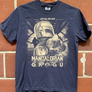 Puede incluir: Camiseta azul marino con un gr&aacute;fico de The Mandalorian y Grogu de Star Wars. El dise&ntilde;o incluye el texto "STAR WARS" encima de la imagen y "THE MANDALORIAN and GROGU" debajo.