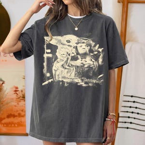 May include: Dark grey t-shirt with a faded graphic of a character from a popular science fiction franchise. The image shows a character holding a baby-like creature. The shirt has a crew neck and short sleeves.