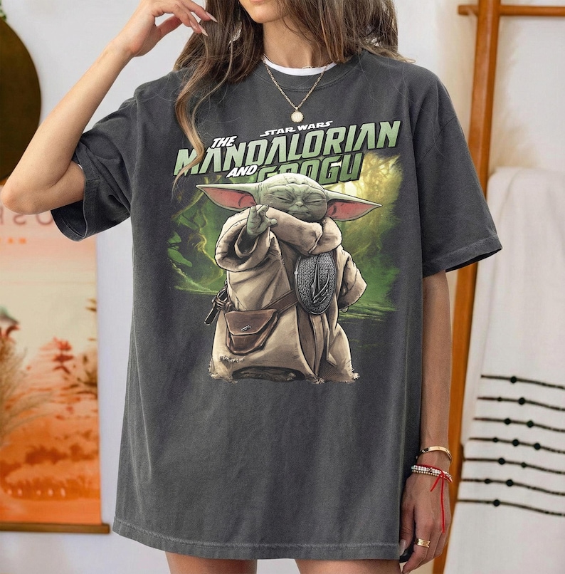 May include: Dark grey t-shirt with a graphic of Grogu from The Mandalorian. The image shows Grogu holding a silver object, with the text "The Mandalorian and Grogu" above. The background is a green and brown landscape.