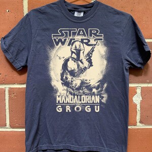 Puede incluir: Camiseta azul marino con un dise&ntilde;o de Star Wars. El dise&ntilde;o presenta a The Mandalorian y Grogu, con el texto "STAR WARS" arriba y "THE MANDALORIAN AND GROGU" abajo. El gr&aacute;fico es en blanco y beige.