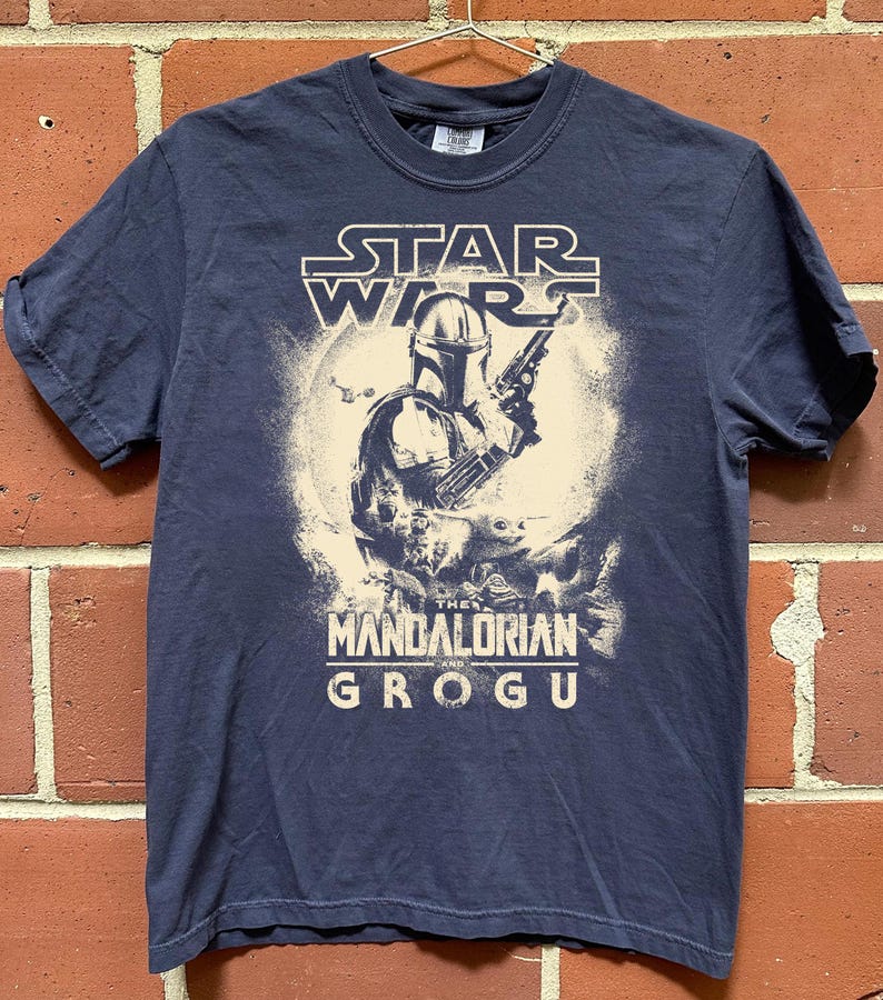 Puede incluir: Camiseta azul marino con un dise&ntilde;o de Star Wars. El dise&ntilde;o presenta a The Mandalorian y Grogu, con el texto "STAR WARS" arriba y "THE MANDALORIAN AND GROGU" abajo. El gr&aacute;fico es en blanco y beige.