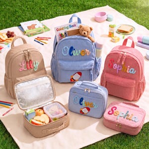 Op de afbeelding: Drie rugzakken en drie bijpassende lunchtassen van corduroy in beige, lichtblauw en roze. Elk item is voorzien van de geborduurde namen Lily, Oliver of Sophia. De blauwe rugzak bevat een teddybeer en raketpatch, de lunchtassen hebben een zilveren isolatie.
