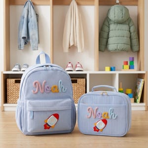 Op de afbeelding: Een lichtblauwe rugzak en bijpassende lunchtas van corduroy. Beide items zijn voorzien van de naam Noah in reli&euml;f met meerkleurige garenletters en een raket-patch. De rugzak heeft een handvat aan de bovenkant en een voorvak met rits.