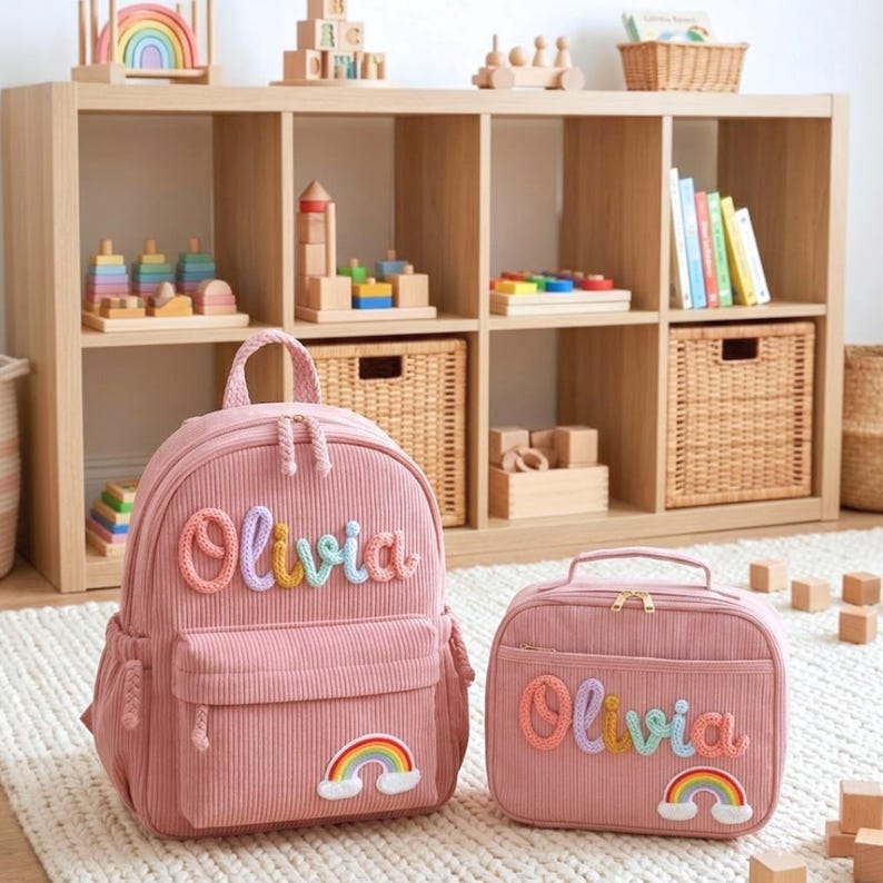 Op de afbeelding: Een roze rugzak van corduroy en een bijpassende lunchtas, beide voorzien van de naam Olivia in veelkleurige, textuurrijke garenletters en een kleine regenboogpatch met witte wolken. De items hebben goudkleurige ritssluitingen.