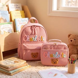 Op de afbeelding: Een roze rugzak en bijpassende lunchtas van corduroy, beide voorzien van de naam Charlotte geborduurd in kleurrijke garenletters en een kleine bruine teddybeer-patch. De items hebben goudkleurige ritsen en een geribbelde textuur.