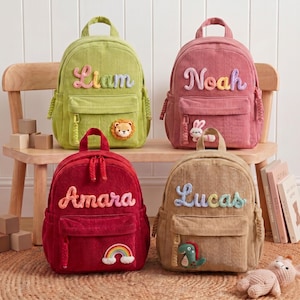 May include: Four corduroy backpacks in lime green, dusty pink, deep red, and tan. Each bag features a top handle, a front zippered pocket, and a personalized name in colorful yarn lettering. Decorative patches include a lion, rabbit, rainbow, and dinosaur.