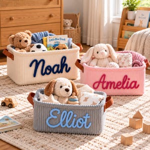May include: Three rectangular storage baskets made of ribbed fabric with brown faux leather handles. The white basket features the name Noah in dark blue, the pink basket features Amelia in dark pink, and the gray basket features Elliot in light blue.