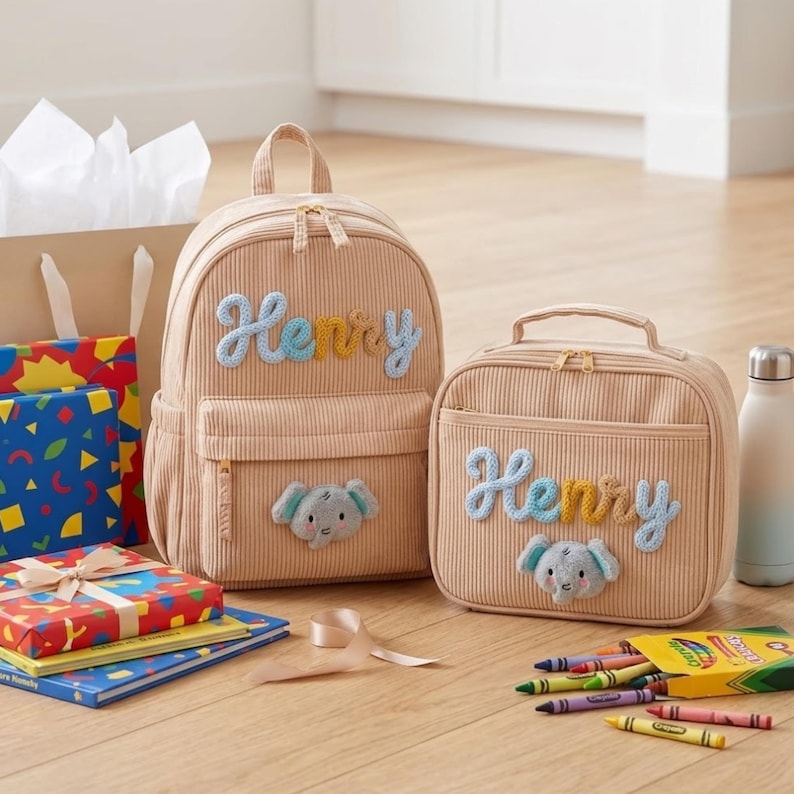 Op de afbeelding: Een beige rugzak van corduroy en een bijpassende lunchtas, beide voorzien van de naam Henry in blauwe en gele garenletters en een kleine olifantenpatch. De items staan naast kleurrijke geschenkdozen en een doosje met kleurpotloden.