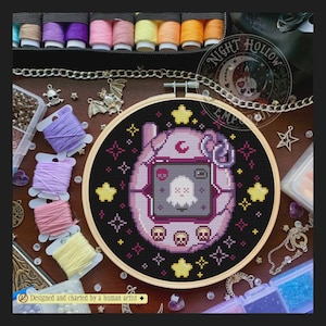 Puede incluir: Un bastidor de bordado circular de madera con un patrón de punto de cruz de un dispositivo de mascota digital de color morado claro sobre fondo negro. La pantalla muestra un fantasma blanco con un ojo en forma de X. Estrellas amarillas y calaveras decoran el diseño.