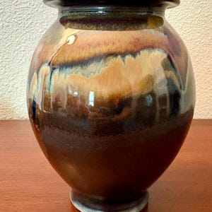 May include: A ceramic vase with a rounded body and a wide, flared rim. The surface features a glossy glaze with blended layers of deep brown, tan, and hints of blue-green that drip down the sides, creating a smooth, multi-toned finish.
