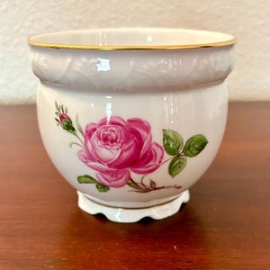 May include: A white porcelain bowl featuring a vibrant pink rose blossom with green leaves and a small bud on the side. The bowl has a scalloped base and a gold-trimmed rim, with a subtle embossed vine pattern encircling the upper section.