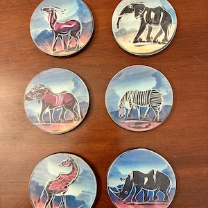 May include: A set of six circular coasters featuring stylized illustrations of African wildlife, including an antelope, elephant, lion, zebra, giraffe, and rhinoceros. Each coaster displays a vibrant sunset-toned background with blue skies.