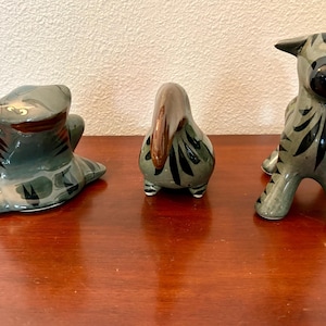 May include: Three ceramic animal figurines with a glossy grey finish and dark black painted stripes. The set includes a crouching frog, a small bird with a brown beak, and a standing zebra-like creature with a rounded black nose.