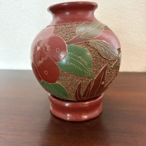 May include: A small, round, reddish-brown vase with a smooth neck and base. The body features a textured, brown, pebbled surface decorated with carved, painted floral motifs in shades of red, green, and brown, resting on a pedestal base.