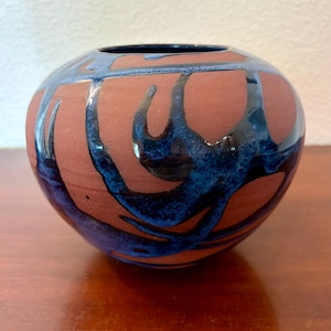 May include: A round, bulbous ceramic vase with a matte terracotta-colored base. The surface features a glossy, dark blue glaze applied in fluid, abstract, dripping patterns that contrast with the unglazed, earthy red clay texture of the vessel.
