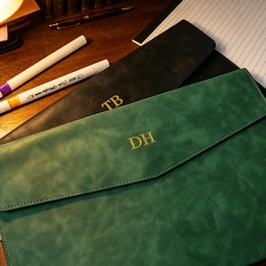 Puede incluir: Portadocumentos de cuero verde y negro con iniciales doradas. El portadocumentos verde tiene las iniciales "DH" y el negro "TB". Se ven dos rotuladores blancos con puntas moradas y naranjas, junto con un bolígrafo y un bloc de notas.