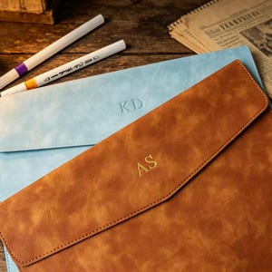 Könnte beinhalten: Hellbraunes Leder-Portfolio mit goldgeprägten Initialen "AS" und hellblaues Portfolio mit "KD". Weiße Marker mit farbigen Spitzen und Text liegen auf einer Holzoberfläche.