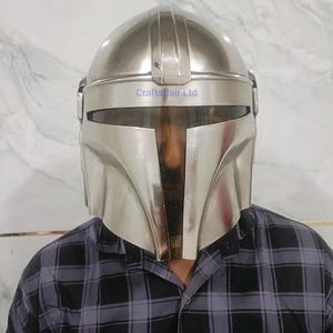 Pu&ograve; includere: Un casco Mandaloriano color argento con una visiera a forma di T. Il casco ha una parte superiore arrotondata e un design angolare distintivo. Il testo "Craftsflair Ltd" &egrave; visibile sul casco. La persona indossa una camicia a quadri neri e grigi.