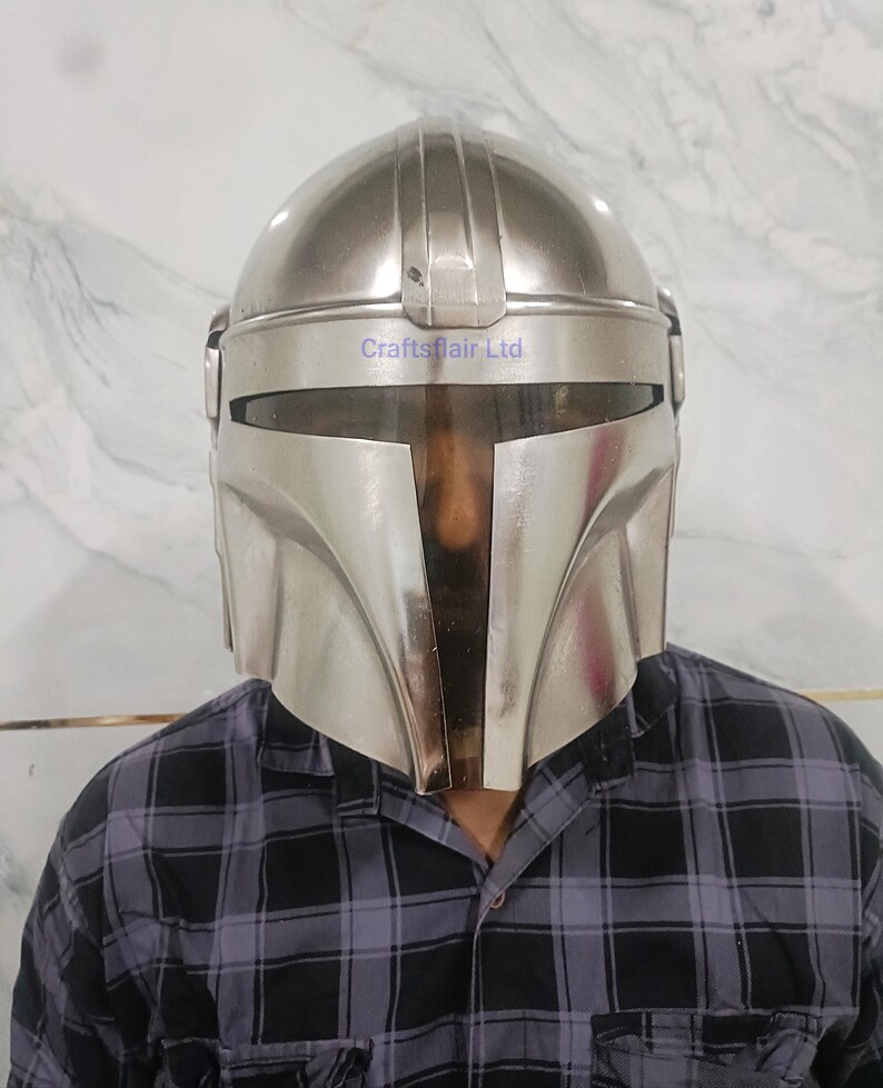 Pu&ograve; includere: Un casco Mandaloriano color argento con una visiera a forma di T. Il casco ha una parte superiore arrotondata e un design angolare distintivo. Il testo "Craftsflair Ltd" &egrave; visibile sul casco. La persona indossa una camicia a quadri neri e grigi.