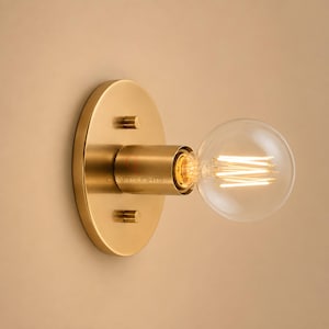 May include: A brass wall sconce featuring a circular mounting plate with two small knobs and a cylindrical socket holding a clear, round Edison-style light bulb with a visible glowing filament. The text CRAFTSLIGHTS is printed on the fixture.