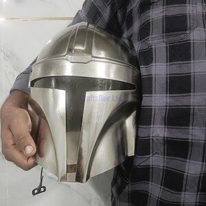 Pu&ograve; includere: Un casco color argento con una visiera a forma di T distintiva, che ricorda un casco Mandaloriano. Il casco &egrave; tenuto da una persona. Il casco ha una finitura metallica liscia e un design dettagliato.