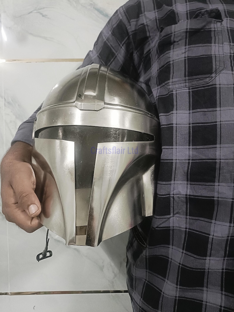 Pu&ograve; includere: Un casco color argento con una visiera a forma di T distintiva, che ricorda un casco Mandaloriano. Il casco &egrave; tenuto da una persona. Il casco ha una finitura metallica liscia e un design dettagliato.