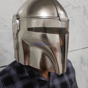 Pu&ograve; includere: Un casco argentato Mandaloriano con visiera e apertura a forma di T. Il casco ha una superficie liscia e riflettente ed &egrave; indossato da una persona con una camicia a quadri. Il casco &egrave; una replica di quello indossato nella serie Star Wars.