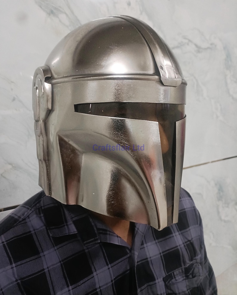 Pu&ograve; includere: Un casco argentato Mandaloriano con visiera e apertura a forma di T. Il casco ha una superficie liscia e riflettente ed &egrave; indossato da una persona con una camicia a quadri. Il casco &egrave; una replica di quello indossato nella serie Star Wars.