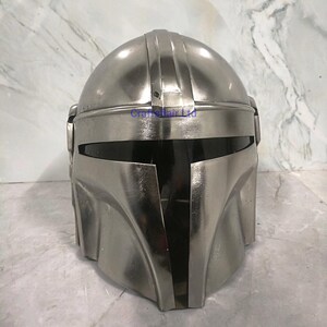 Pu&ograve; includere: Un casco color argento con visiera e parte superiore arrotondata. Il casco ha una finitura metallica e sembra essere realizzato in un materiale resistente. Il design include creste verticali ed elementi angolari, suggerendo un tema futuristico o fantascientifico.