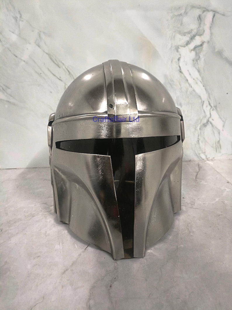Pu&ograve; includere: Un casco color argento con visiera e parte superiore arrotondata. Il casco ha una finitura metallica e sembra essere realizzato in un materiale resistente. Il design include creste verticali ed elementi angolari, suggerendo un tema futuristico o fantascientifico.