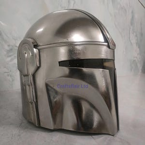 Pu&ograve; includere: Un casco argentato con visiera e design distintivo. Il casco ha una superficie liscia e riflettente e presenta una prominente cresta sopraccigliare e dettagli laterali. Il testo "Craftsflair Ltd" &egrave; visibile in basso a destra del casco.