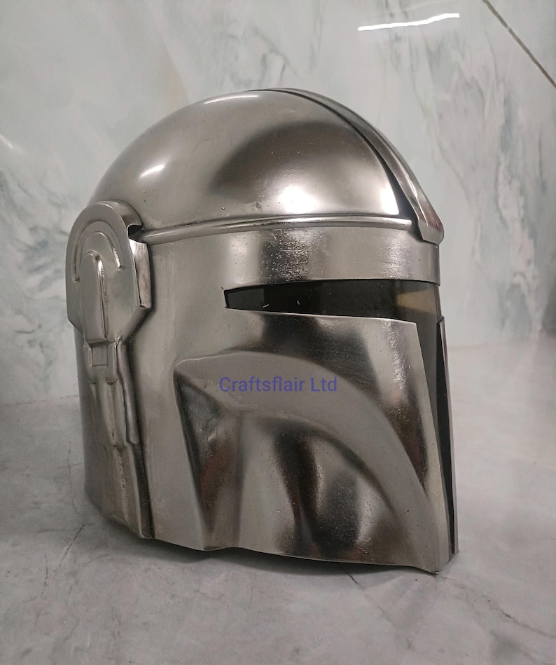 Pu&ograve; includere: Un casco argentato con visiera e design distintivo. Il casco ha una superficie liscia e riflettente e presenta una prominente cresta sopraccigliare e dettagli laterali. Il testo "Craftsflair Ltd" &egrave; visibile in basso a destra del casco.
