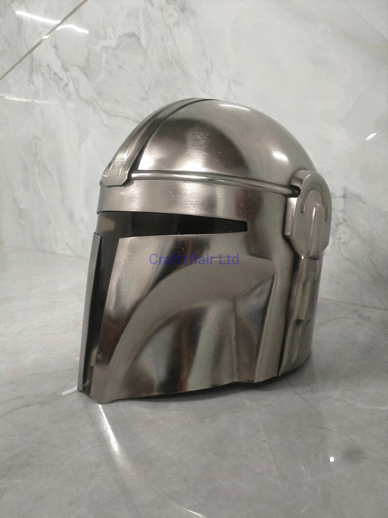 Pu&ograve; includere: Un casco Mandaloriano color argento, una replica del franchise di Star Wars. Il casco presenta una visiera a forma di T e una parte superiore arrotondata. La superficie riflette la luce, conferendogli un aspetto metallico.