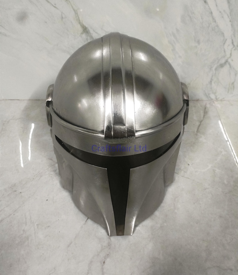 Pu&ograve; includere: Un casco color argento con una visiera a forma di T. Il casco ha una parte superiore arrotondata e una finitura metallica liscia. Il design include creste verticali e una prominente linea delle sopracciglia.