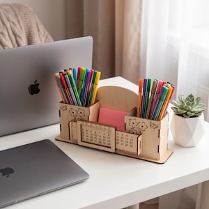 Peut inclure: Un organisateur de bureau en bois clair doté de deux porte-stylos carrés avec des motifs de fleurs et de papillons découpés au laser. La section centrale contient un calendrier perpétuel coulissant et un bloc de notes adhésives roses.