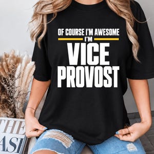 Pode incluir: Uma t-shirt preta de manga curta com o texto branco "OF COURSE I'M AWESOME I'M VICE PROVOST" e uma linha horizontal amarela separando as duas primeiras linhas de texto. A t-shirt é combinada com calções de ganga azul-claros desgastados.