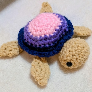 May include: A crocheted turtle toy featuring a tan head and four flippers, with a multi-colored shell consisting of a central pink circle surrounded by a purple ring and a dark blue outer border, finished with a small black bead for the eye.