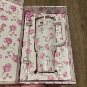 Op de afbeelding: Een witte Stanley-beker met een roze rozenpatroon, een witte handgreep en een doorzichtig rietje. De beker zit in een bijpassende bloemenbox. Op de beker staat de tekst "STANLEY" en "LOVES SHADY LACY".