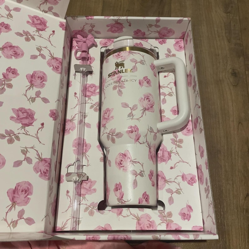 Op de afbeelding: Een witte Stanley-beker met een roze rozenpatroon, een witte handgreep en een doorzichtig rietje. De beker zit in een bijpassende bloemenbox. Op de beker staat de tekst "STANLEY" en "LOVES SHADY LACY".